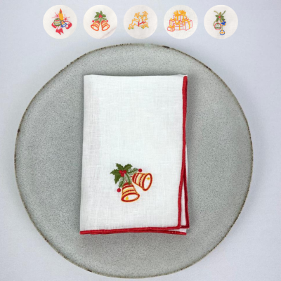 Guardanapo branco dobrado com bordado de sinos e folhas sobre prato cinza.