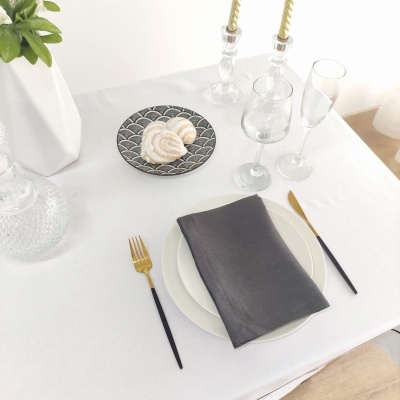 Mesa posta com prato branco, guardanapo cinzento escuro, talheres dourados e pretos, taças, velas em castiçais e prato com biscoitos.