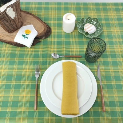 Mesa posta com toalha xadrez, prato branco, guardanapo amarelo, talheres, copo verde e decoração com vela e pãozinho
