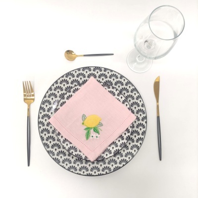 Conjunto de mesa com prato padrão preto e branco, guardanapo rosa bordado, talheres dourados e pretos, colher pequena dourada e copo de vidro transparente.