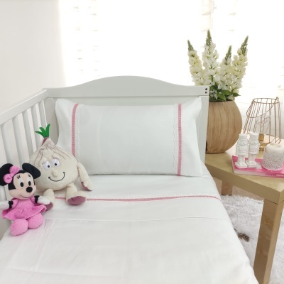 Cama de bebé branca com lençol e fronha brancos com detalhes rosa, peluches Minnie e personagem branca, mesa ao lado com flores e decoração