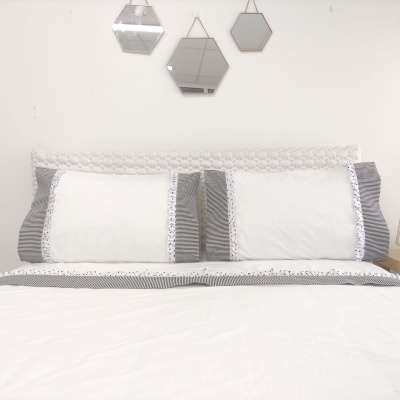 Almofadas brancas com bordas cinzentas e renda numa cama com cabeceira branca e espelhos geométricos na parede