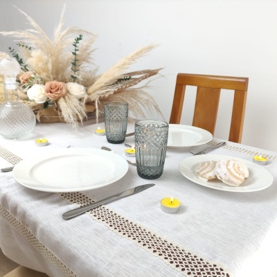 Mesa posta com toalha branca, pratos brancos, copos cinza texturizados, faca, velas amarelas e arranjo de flores secas e rosas