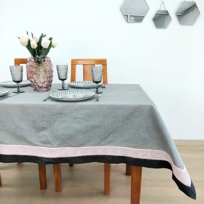 Toalha de mesa cinzenta com padrão na borda, mesa de madeira com vasos, pratos e copos, fundo branco com espelhos.