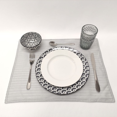 Conjunto de loiça com prato e taça decorados, talheres em metal e copo, sobre toalhete bege.