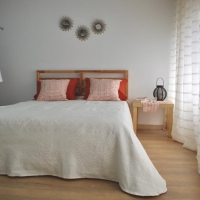 Quarto com cama de madeira e colcha bege clara, duas almofadas castanho-avermelhadas e cortinas brancas translúcidas.