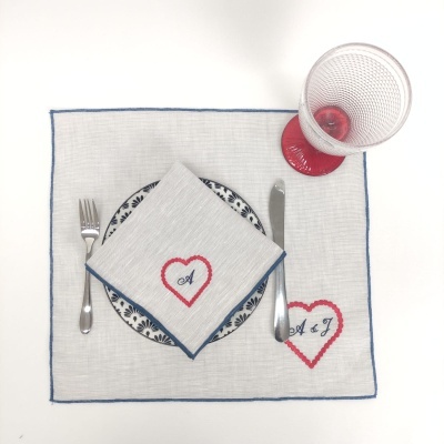 Mesa posta com individual branco bordado, guardanapo, prato azul e branco, talheres e copo com base vermelha.