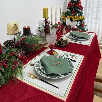 Mesa de Natal decorada com toalha vermelha, velas, pinhas, pratos e guardanapos verdes bordados, talheres dourados, copos de cristal e árvore de Natal ao fundo