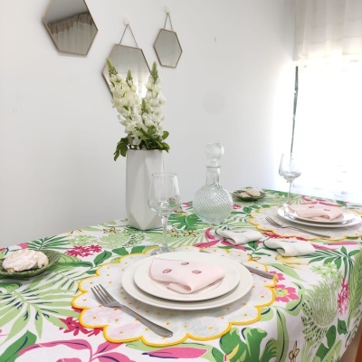 Mesa posta com toalha de padrão floral colorido, pratos brancos, guardanapos cor-de-rosa, copos e garrafa de vidro, vaso branco com flores e três espelhos geométricos na parede.