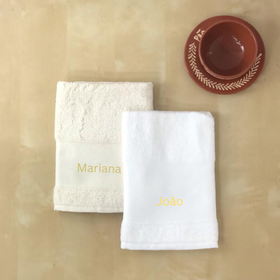 Toalhas de banho creme e branca com nomes bordados ao lado de prato e recipiente vermelhos