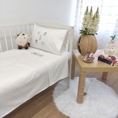 Cama branca com roupa de cama e desenhos, mesa de madeira com relógio, vaso com flores e peluches