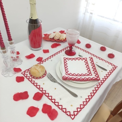 Mesa posta com jogo americano e guardanapo bordados em branco e vermelho, pão, talheres, taça, garrafa, velas e pétalas vermelhas.