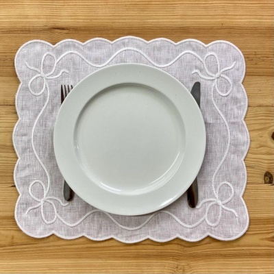 Lugar americano branco bordado com prato branco e talheres numa mesa de madeira.