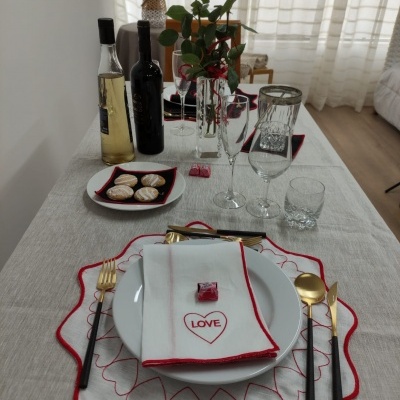 Mesa posta com toalha bege, pratos brancos, guardanapo com LOVE, talheres dourados e pretos, taças, garrafas, biscoitos e rosas vermelhas