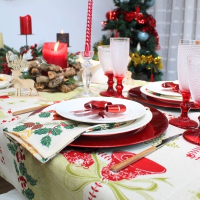 Mesa de Natal com pratos brancos em sousplats vermelhos, guardanapo decorado, taças vermelhas e transparentes, velas vermelhas acesas e árvore de Natal ao fundo