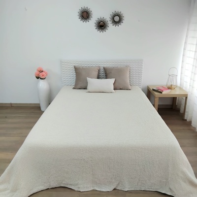 Cama de casal com colcha bege clara e almofadas castanhas e bege, parede branca, vaso com flores e mesa de cabeceira