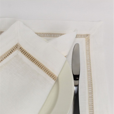 Guardanapo branco com bordado bege junto a prato branco e faca prateada sobre toalha de mesa branca