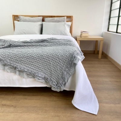 Cama com lençóis brancos e manta cinza trançada num quarto com mesa de cabeceira e livros