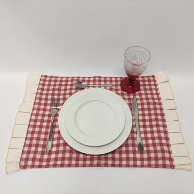 Posto de mesa com prato branco, talheres, copo vermelho e toalha xadrez vermelha e branca.