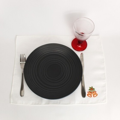 Conjunto de jantar com prato preto circular, talheres metálicos, copo com base vermelha e individual branco com bordado de sinos.
