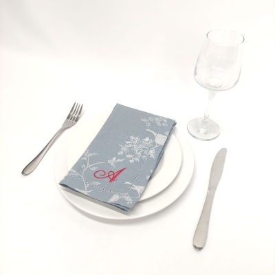 Conjunto de mesa com garfo, faca, prato branco, guardanapo azul com padrão floral, letra A vermelha e copo de vidro