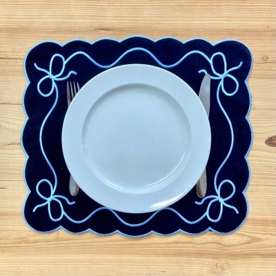 Lugar americano azul escuro com bordados claros, prato branco, garfo e faca sobre mesa de madeira