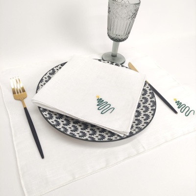 Jogo americano branco com bordado de árvore de Natal, prato branco e azul, talheres dourados e copo de vidro.