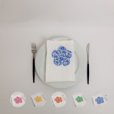 Guardanapo branco com padrão floral azul sobre prato branco com talheres e amostras de padrões florais coloridos.