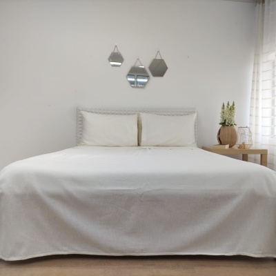 Cama de casal com colcha branca e cabeceira cinzenta, mesa de cabeceira com plantas e candeeiro, espelhos geométricos na parede