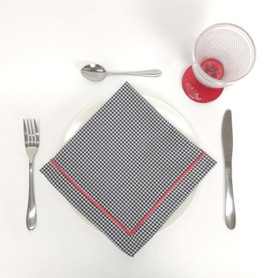 Mesa posta com prato branco, guardanapo xadrez preto e branco com borda vermelha, talheres metálicos e copo com base vermelha.