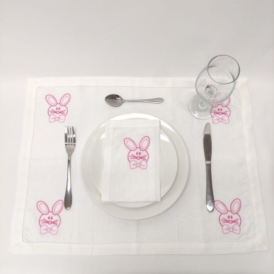 Individual branco com bordados de coelhinhos cor-de-rosa, guardanapo, talheres e taça num fundo branco