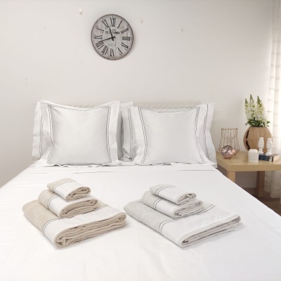 Cama com roupa de cama branca, toalhas dobradas em bege e branco, relógio de parede, mesa de cabeceira com objetos