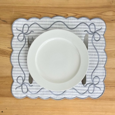 Jogo americano retangular de tecido cinzento com riscas e laços, prato branco e talheres em mesa de madeira