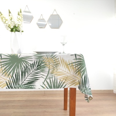 Toalha de mesa com folhas tropicais verde e amarelas sobre fundo branco, mesa de madeira, espelhos pequenos e flores brancas num vaso