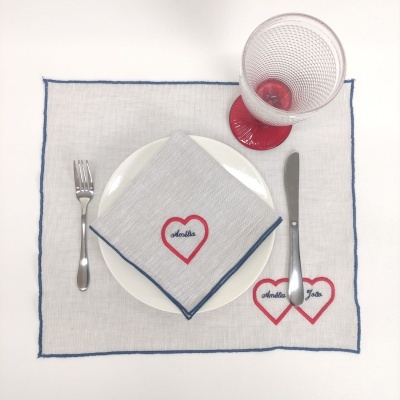 Conjunto de refeição com individual branco, prato, talheres metálicos, guardanapo cinza e copo com pé vermelho