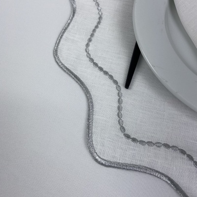 Detalhe de toalha de mesa branca com bordado prateado ao lado de prato e guardanapo