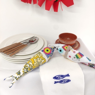 Mesa posta com dois peixes de tecido coloridos, pratos brancos, talheres com cabo de madeira, toalha bordada e chávena de barro.