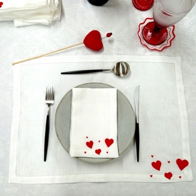 Conjunto de jantar com prato cinzento, talheres pretos, guardanapo com corações vermelhos, toalhete branco com corações vermelhos, copo e decoração vermelha.