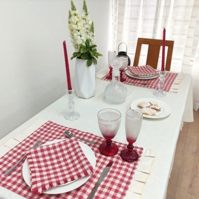 Mesa de jantar posta com toalha branca, guardanapos e individuais xadrez vermelho e branco, pratos e talheres, taças e velas vermelhas, vaso com flores brancas.