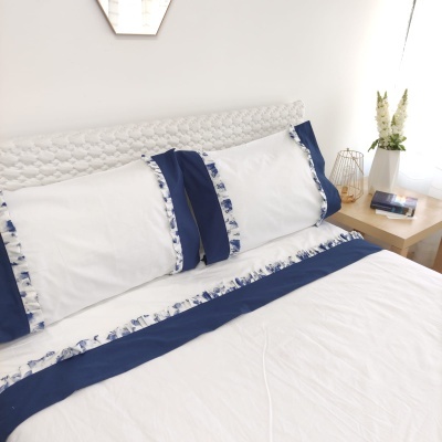 Capa de almofada e sábana brancas com detalhes em azul e padrão floral azul e branco numa cama com cabeceira trançada e mesa de cabeceira com decoração.