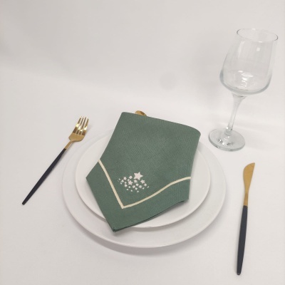 Conjunto de jantar com prato branco, guardanapo verde bordado, talheres dourados e pretos e copo de vinho de vidro