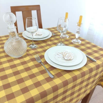 Mesa posta com toalha xadrez amarela e castanha, pratos, copos de vinho, velas e licoreiro em vidro texturizado