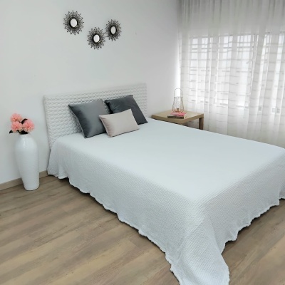 Cama de casal com roupa de cama branca, almofadas cinza e bege, mesa de cabeceira com abajur, vaso com flores rosas, espelhos redondos na parede e janela com cortinas brancas.