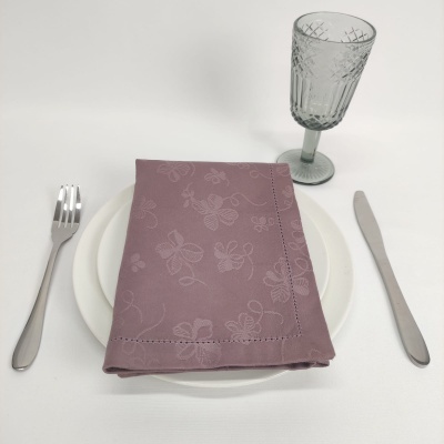 Mesa posta com garfo, faca, prato branco, guardanapo lilás com padrão de abelhas e copo de vidro cinzento