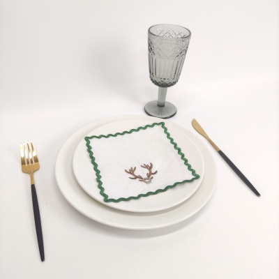 Conjunto de pratos brancos, guardanapo bordado com verde, talheres preto e dourado e copo de vidro cinzento