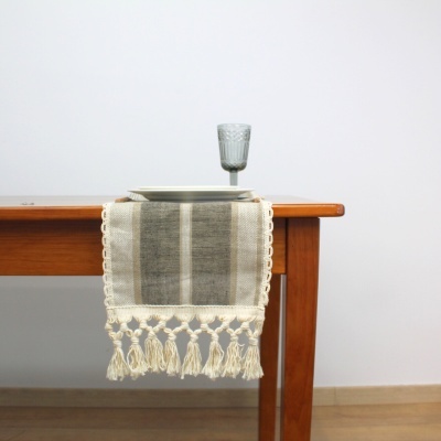 Toalha de mesa bege com franjas numa mesa de madeira com prato branco e copo cinzento