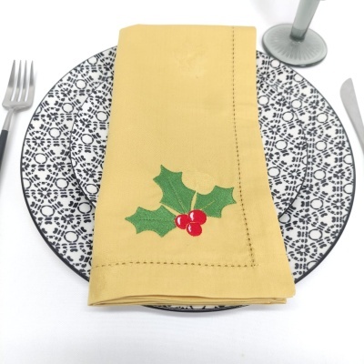 Prato com padrão preto e branco, guardanapo amarelo com bordado de folhas e frutos vermelhos, talheres e base de copo.