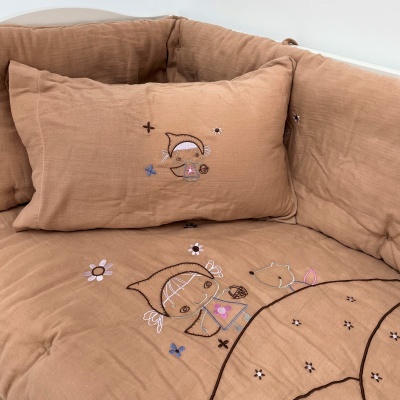 Almofada e edredão castanhos claros com bordados de bonecos e flores coloridas.