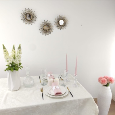 Mesa de jantar decorada com toalha branca, arranjos florais, pratos e guardanapos rosa, talheres dourados, castiçais transparentes e espelhos decorativos no fundo branco.