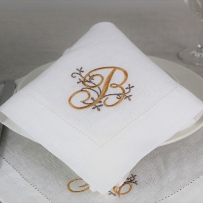 Guardanapo branco bordado com letra B dourada sobre toalha cinzenta e talheres